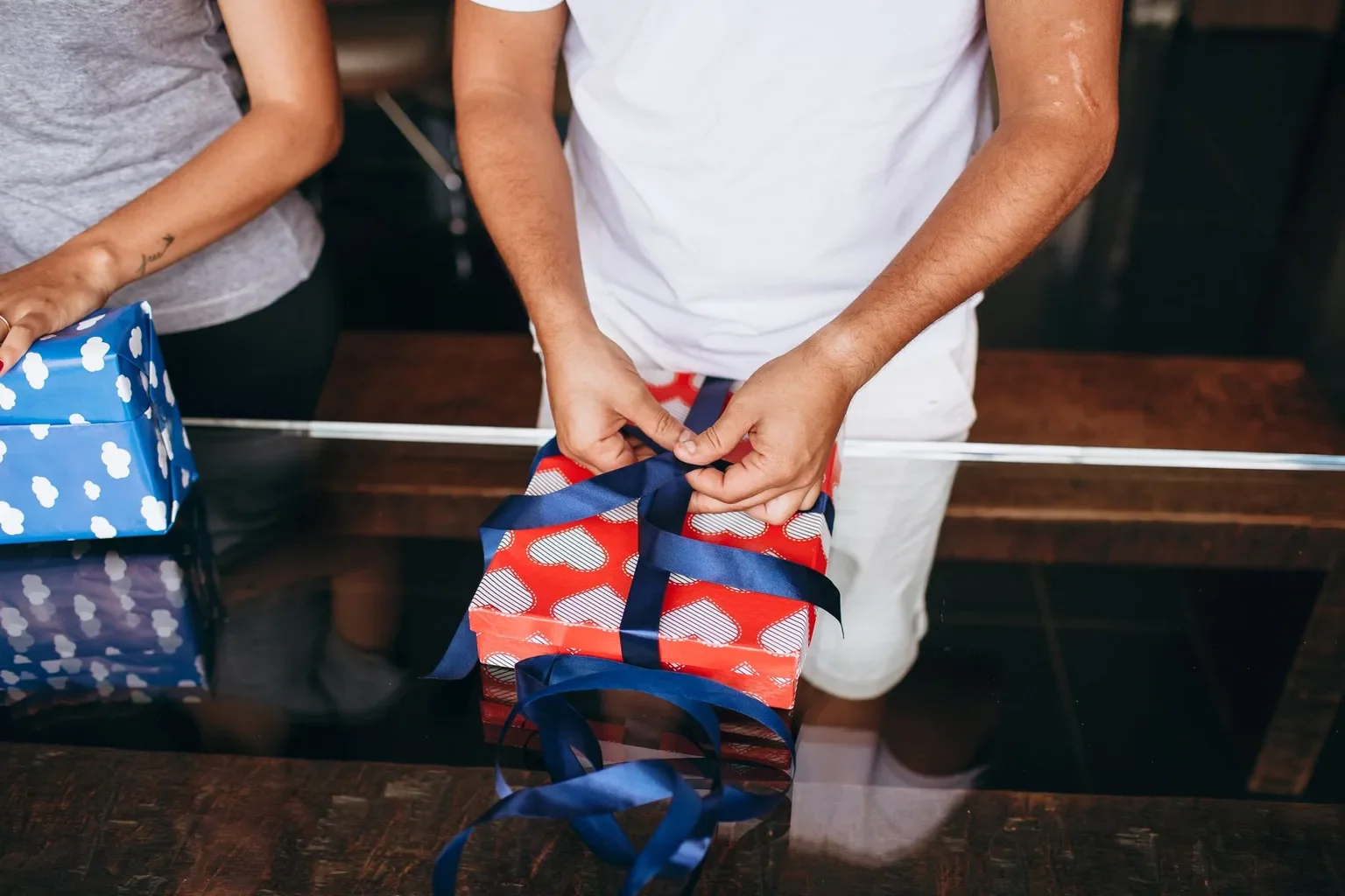 A person tying a blue ribbon around a wrapped gift on a glass table