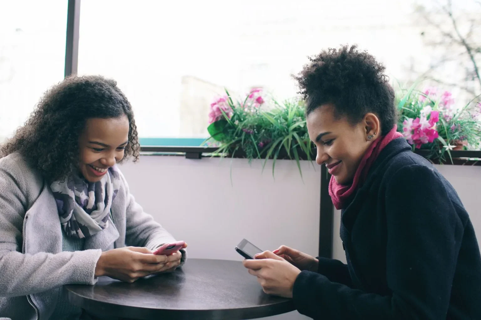 Two friends smiling while using their phones at a table